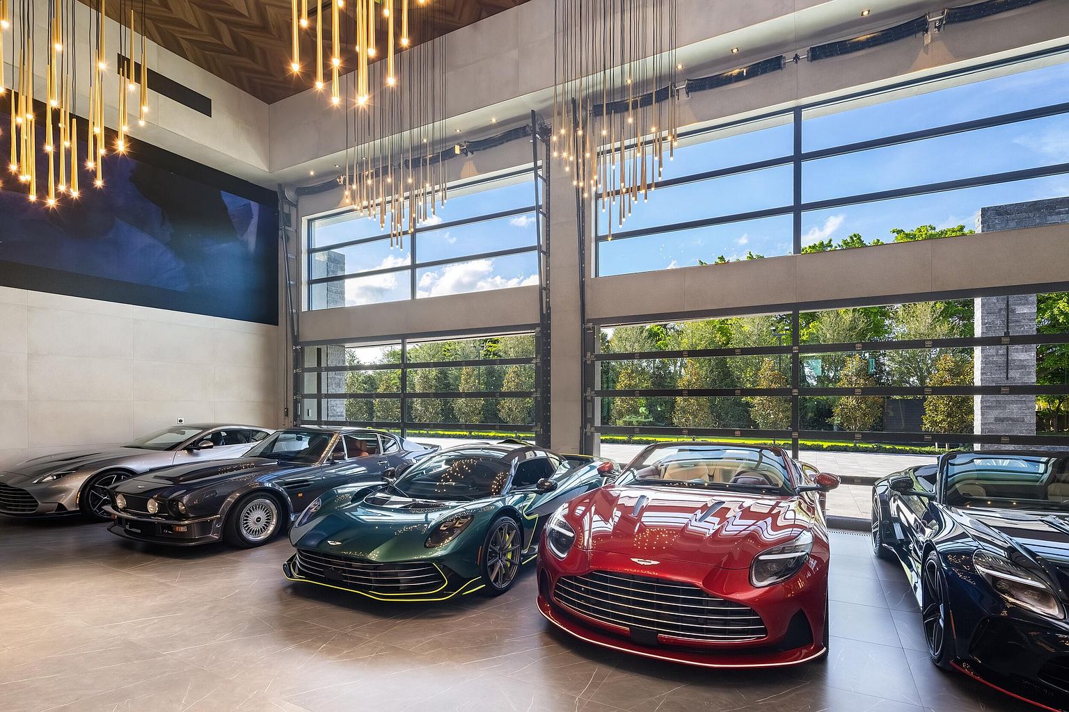 This expansive, high-end luxury garage showcases a collection of premium sports cars arranged on a polished tile floor. The space features dramatic, multi-tiered pendant lighting and massive, industrial-style glass garage doors that provide a clear view of the lush greenery outside. The architectural design emphasizes a sleek, modern aesthetic, creating a gallery-like atmosphere for the vehicles.
