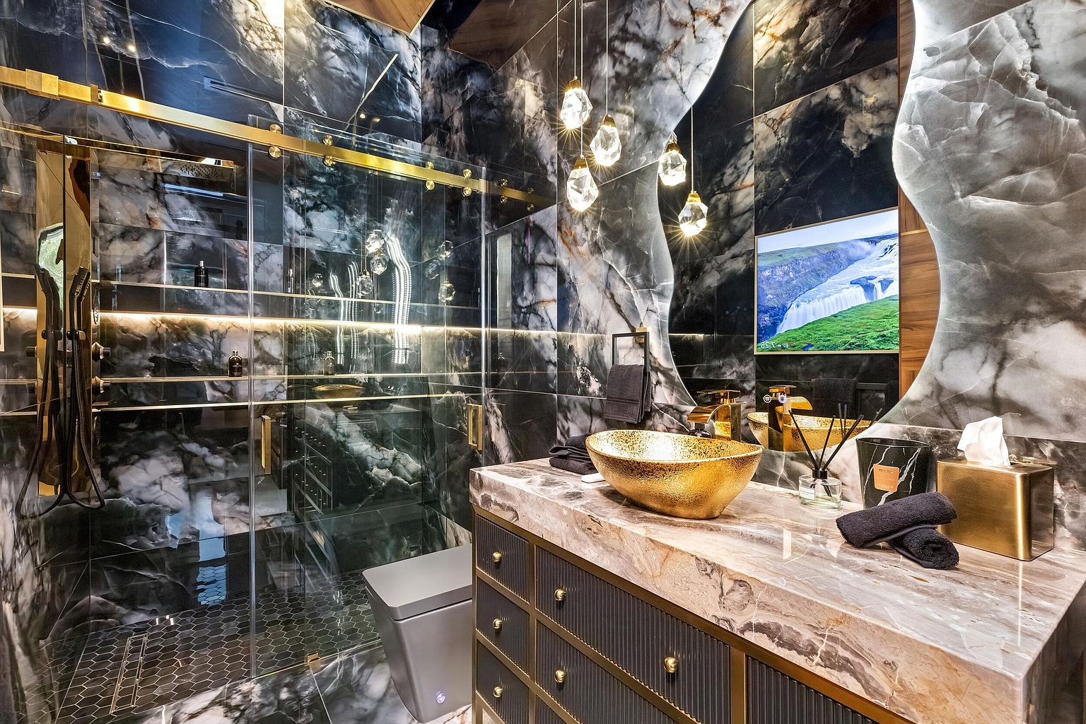 This luxurious bathroom features dramatic dark, veined stone wall tiles and a matching stone vanity countertop. A striking gold-toned vessel sink sits atop a dark, textured vanity cabinet, complemented by a large, uniquely shaped mirror and elegant pendant lighting. The space includes a glass-enclosed shower area with gold hardware and a modern toilet, creating a sophisticated and high-end aesthetic.
