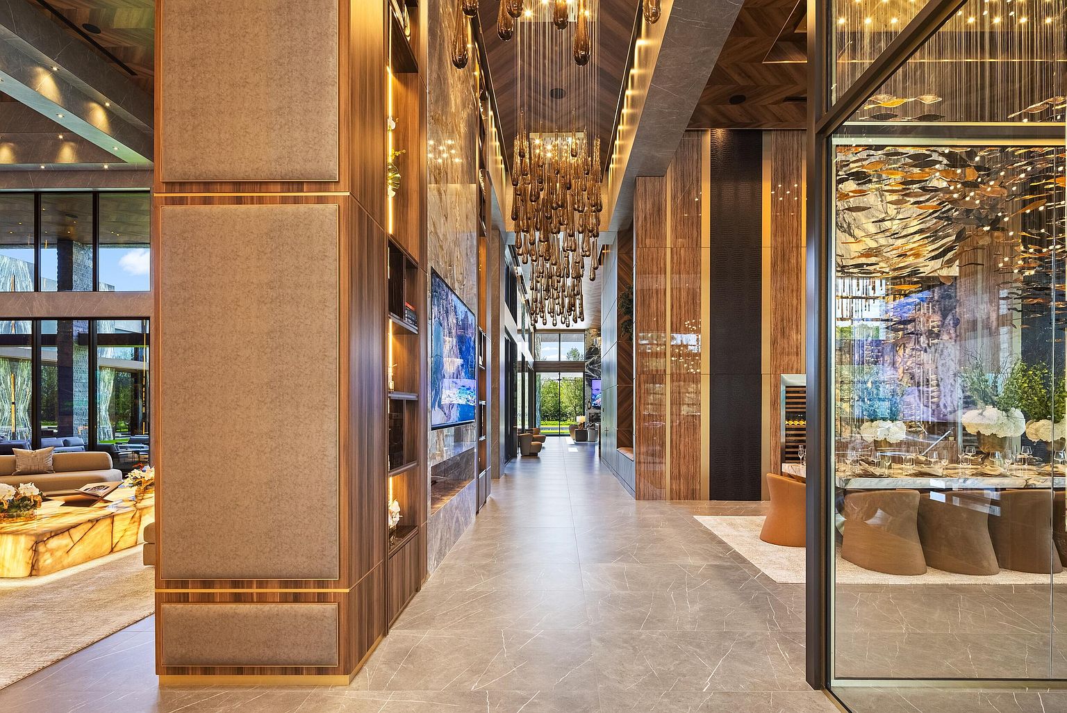This grand hallway serves as a central artery, connecting various living spaces with a sophisticated, high-end aesthetic. A dramatic, multi-tiered chandelier hangs from the ceiling, casting a warm glow over the polished stone flooring and the wood-paneled wall featuring integrated shelving and a television. To the right, a glass-enclosed dining area offers a glimpse of an elaborate art installation, while the perspective draws the eye through the open-concept layout toward the natural light at the far end of the home.