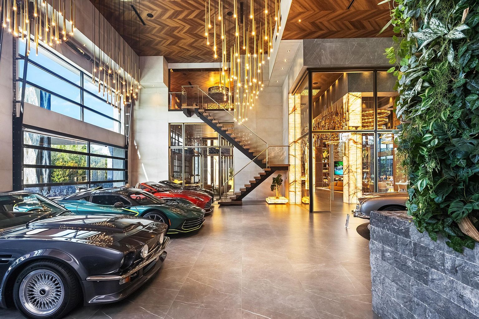 This expansive, high-end garage functions as a luxury showroom, featuring a polished tile floor and a dramatic, multi-tiered chandelier installation hanging from a herringbone-patterned wood ceiling. The space houses a collection of premium vehicles, with large glass garage doors on the left and a staircase leading to an upper mezzanine level. A vertical garden wall adds a touch of nature to the sophisticated, industrial-modern aesthetic.