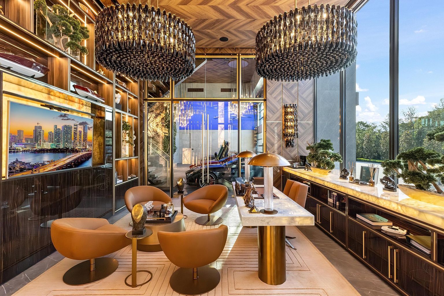 This sophisticated home office features a luxurious, modern aesthetic with a central desk topped with stone and supported by a metallic pedestal. The room is furnished with four tan swivel chairs arranged around a circular coffee table, complemented by extensive dark wood cabinetry and integrated shelving. Large glass walls provide a clear view into an adjacent garage space, while floor-to-ceiling windows on the right offer abundant natural light and a view of the outdoors.