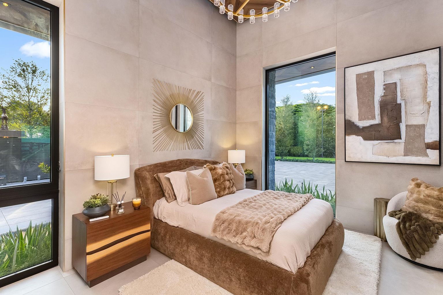 This modern bedroom features a plush, brown upholstered bed frame accented with textured throw pillows and a soft faux-fur throw. The room is framed by large windows that provide abundant natural light and views of the surrounding greenery, while a sunburst-style mirror serves as a focal point above the bed. A sleek wooden nightstand and a contemporary armchair complete the space, which is finished with neutral, large-format wall tiles and a high-end chandelier.