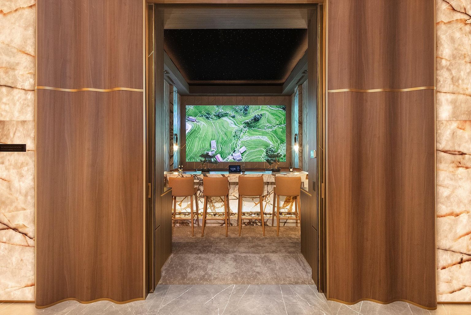 This sophisticated screening room features a central bar area with four leather-upholstered stools set against a backlit stone counter. The room is framed by rich wood-paneled walls with gold accents and a dramatic ceiling featuring a starry night effect. The perspective is centered, looking through a grand doorway into the intimate, high-end lounge space.