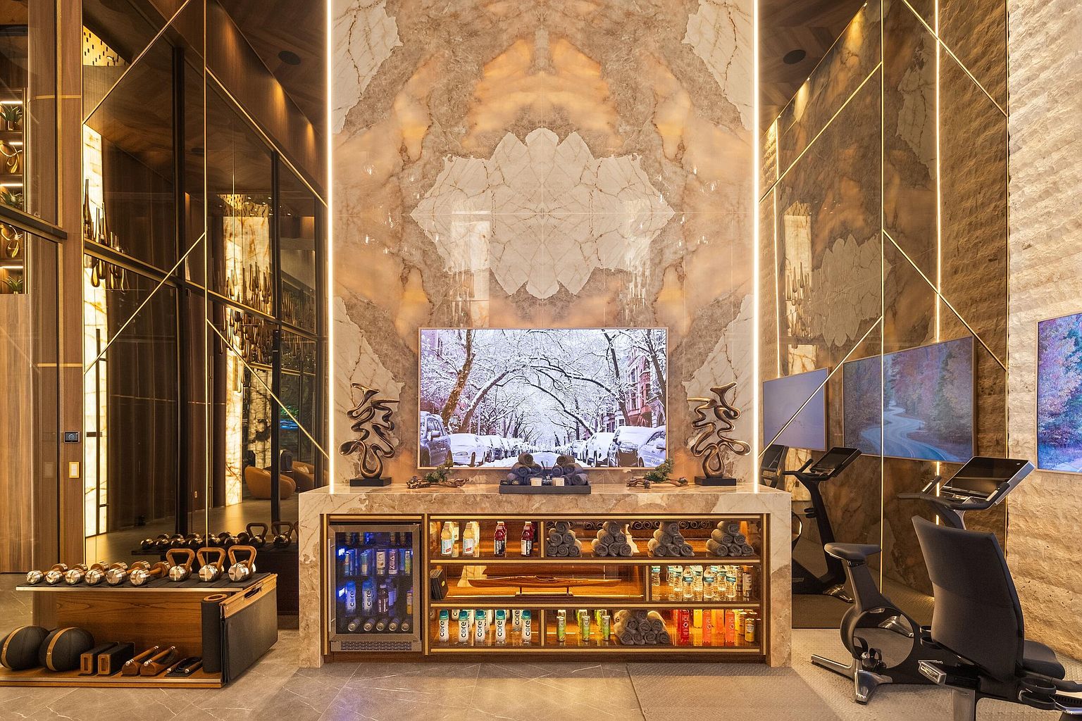 This luxurious home gym features a central stone-clad console with a built-in beverage refrigerator and open shelving stocked with towels and bottled water. A large television is mounted above the console, flanked by decorative metal sculptures, all set against a dramatic, book-matched stone wall with integrated vertical lighting. The space is equipped with professional-grade exercise equipment, including stationary bikes and a rack of dumbbells, creating a high-end, spa-like fitness environment.