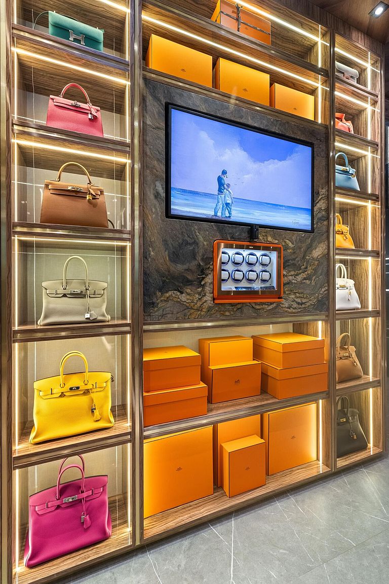 This luxurious walk-in closet features custom wood shelving with integrated LED lighting, designed to showcase a high-end collection of designer handbags and orange storage boxes. A central stone-clad wall serves as a focal point, housing a flat-screen television and a small, illuminated display case. The perspective is a straight-on, eye-level shot that emphasizes the meticulous organization and sophisticated aesthetic of the space.