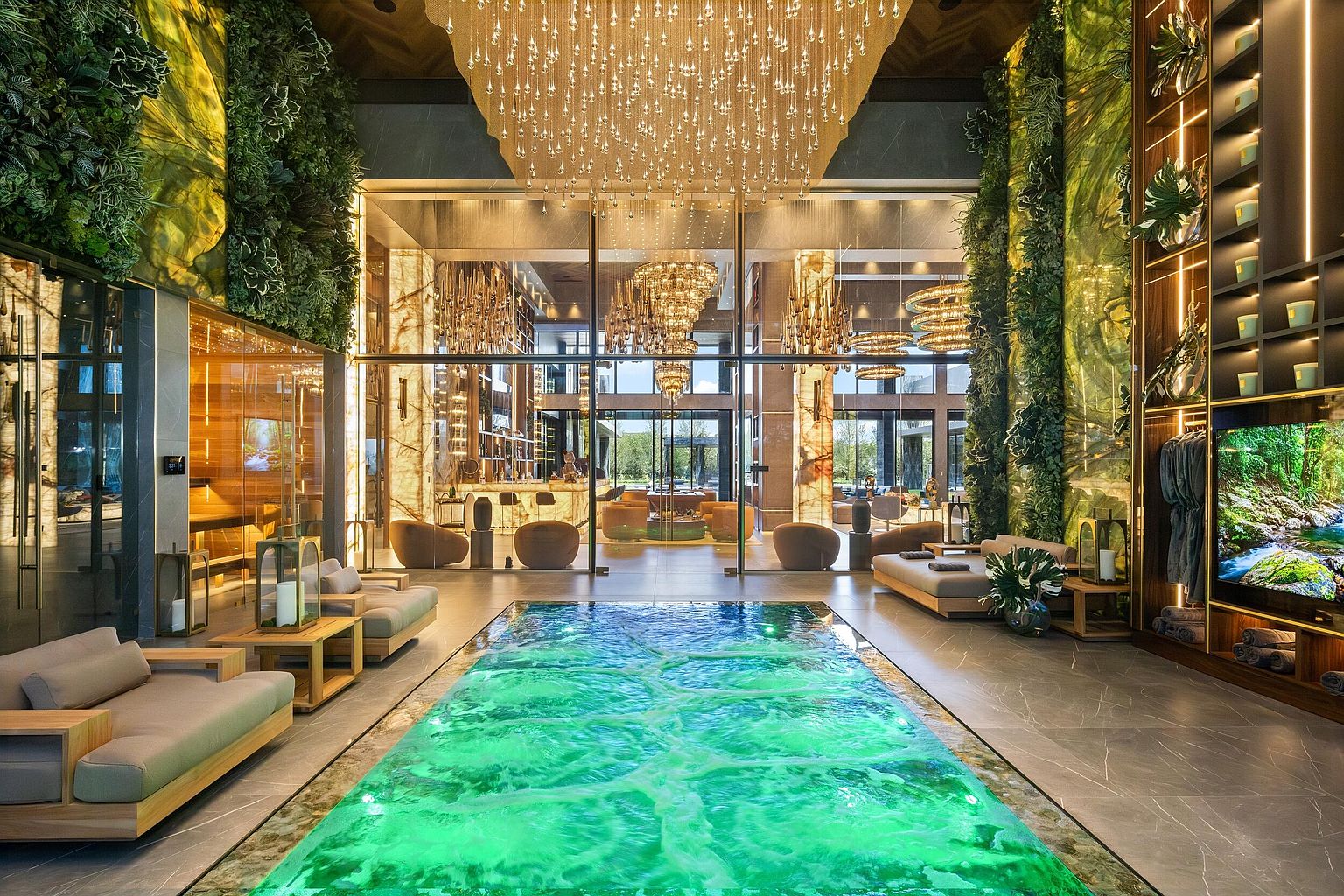 This luxurious indoor spa area features a vibrant, illuminated green pool as the central focal point, surrounded by sleek stone flooring and comfortable lounge seating. The space is framed by lush vertical gardens on either side and a massive, intricate chandelier hanging from the ceiling, while large glass panels provide a clear view into an adjacent lounge area. The overall aesthetic is one of high-end, resort-style opulence, blending natural elements with sophisticated modern design.