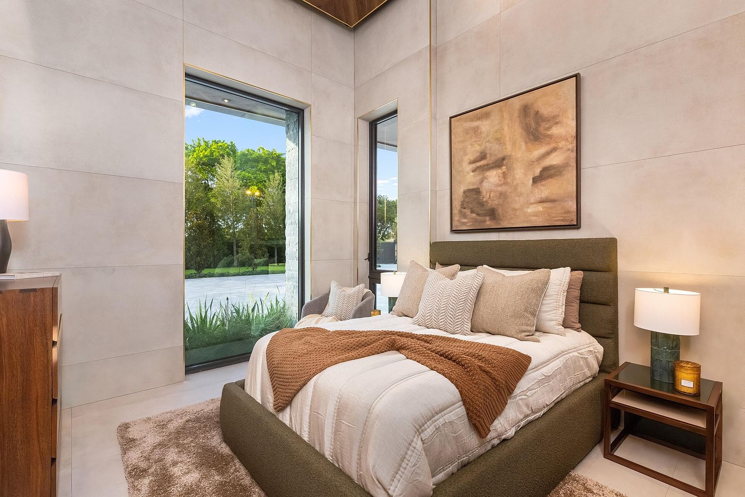 This contemporary bedroom features a textured olive-green upholstered bed frame accented with neutral-toned pillows and a warm, rust-colored throw blanket. Large, floor-to-ceiling windows provide an expansive view of the lush green exterior, while the room is finished with light-toned wall panels and a piece of abstract art above the headboard. The space is illuminated by soft bedside lamps, creating a serene and sophisticated atmosphere.