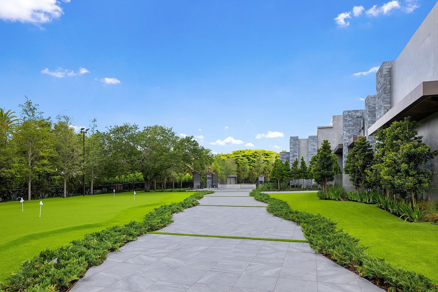 This expansive outdoor space features a meticulously manicured putting green on the left, complete with multiple flag pins, transitioning into a wide, modern stone-paved walkway. The path is bordered by lush, low-lying greenery and leads toward a gated entrance in the distance, flanked by a contemporary building with a textured stone facade. The scene is captured from a ground-level perspective, emphasizing the clean lines, vibrant landscaping, and bright, sunny atmosphere of the property.