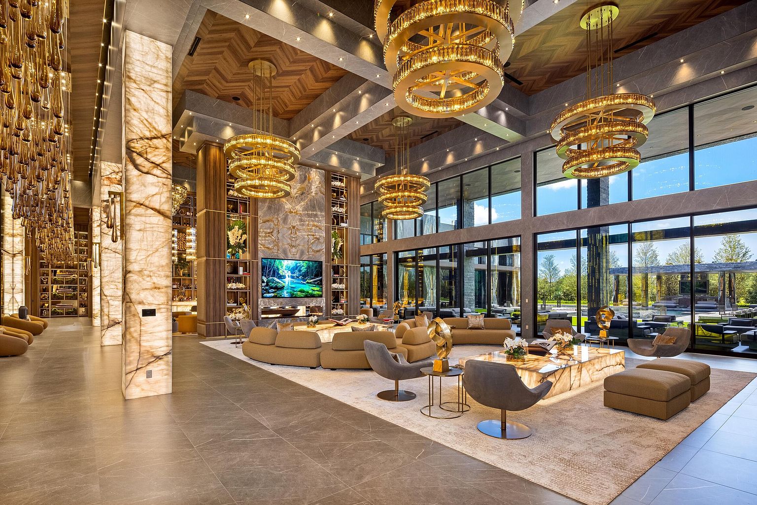 This expansive, high-ceilinged living room showcases a luxurious open-concept design with a massive stone-clad chimney breast serving as a central focal point. The space is illuminated by multiple tiered gold chandeliers and features large windows that provide a seamless connection to the outdoor landscape. A curved sectional sofa and a large stone-topped coffee table anchor the seating area, while a backlit stone pillar adds a warm, dramatic glow to the sophisticated interior.