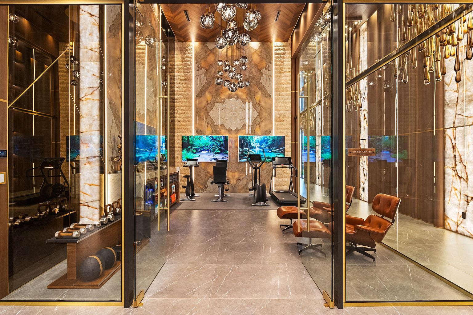 This luxurious, high-end fitness center features a sophisticated design with warm, backlit stone wall panels and a dark wood-paneled ceiling. The space is equipped with professional-grade exercise machines, including treadmills and stationary bikes, positioned in front of large digital displays. A glass-enclosed entrance with gold-toned framing leads into the room, which also includes a dedicated weight training area with dumbbells and leather lounge chairs for relaxation.