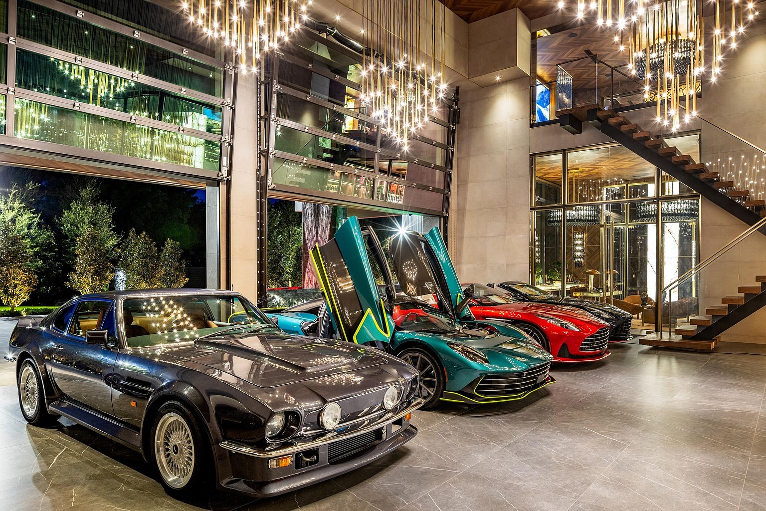 This high-end, contemporary garage space functions as a luxury showroom for a collection of premium vehicles, including a classic coupe and modern sports cars with gull-wing doors. The space features large, retractable glass walls that open to the outdoors, polished stone flooring, and dramatic, multi-tiered crystal chandeliers. A floating staircase with glass railings leads to an upper level, emphasizing the architectural sophistication and grand scale of this automotive display area.