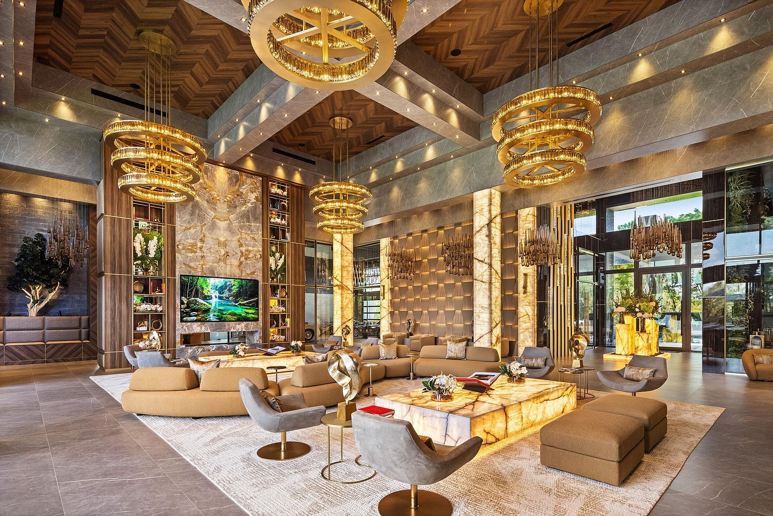 This expansive, high-ceilinged lobby or clubhouse lounge features a sophisticated design with a warm, neutral color palette and intricate wood-paneled ceilings. The space is anchored by a large, illuminated stone coffee table and multiple seating areas with plush, curved sofas and modern armchairs. Multiple tiered gold-toned chandeliers provide dramatic lighting, while a large television is mounted on a prominent stone-clad wall, creating an inviting and luxurious atmosphere.