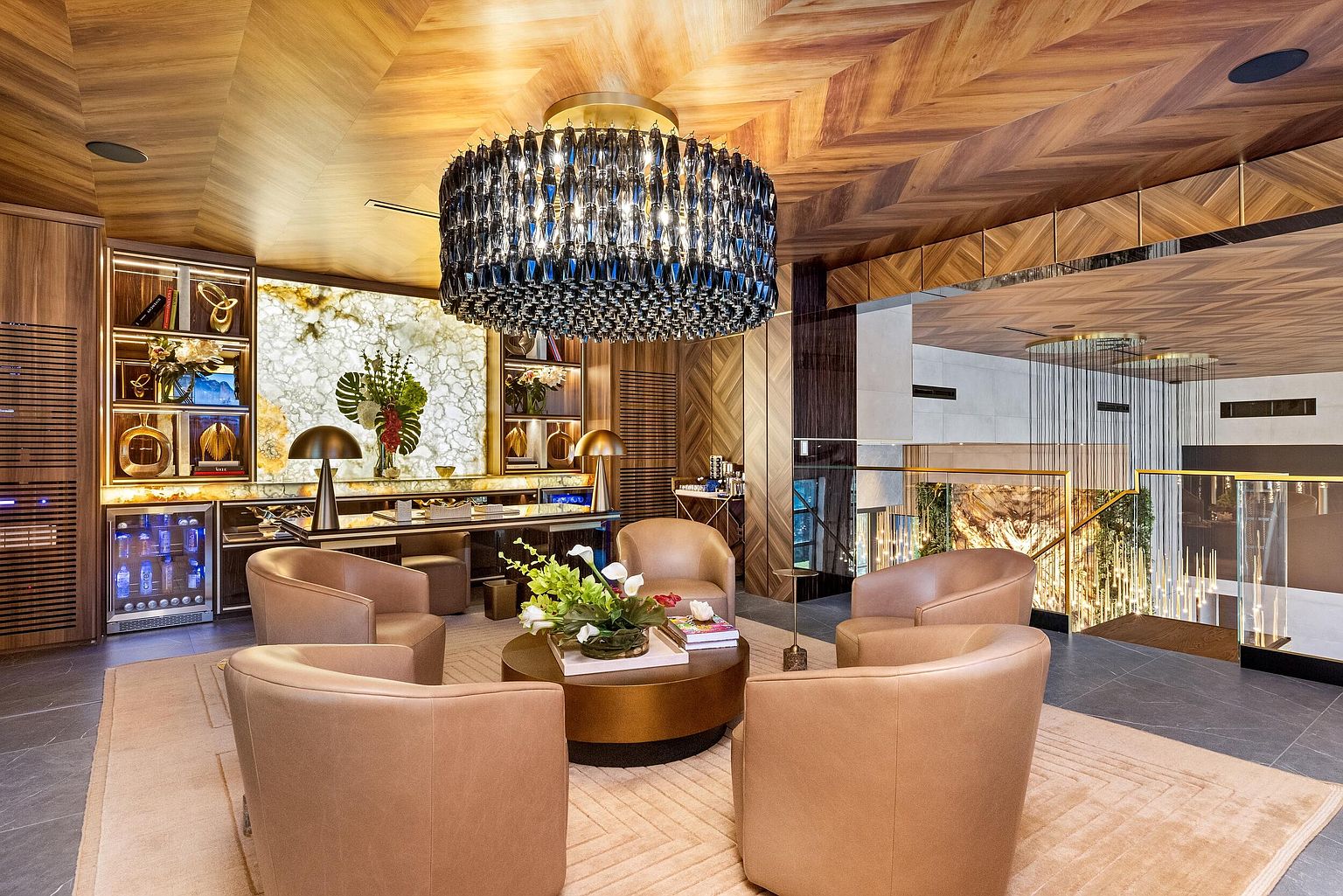 This sophisticated lounge or office space features a central seating area with four tan leather armchairs arranged around a circular coffee table. The room is defined by a striking chevron-patterned wood ceiling and a backlit stone wall behind a desk, creating a luxurious and warm atmosphere. A large, dark crystal chandelier serves as a dramatic focal point, while a glass-fronted beverage cooler and open shelving add functional elegance to the space.