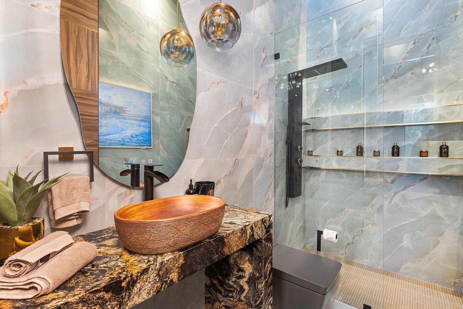 This modern bathroom features a striking vanity topped with a dark, heavily veined stone slab and a carved wooden vessel sink. A large, uniquely shaped mirror with a wood-accented frame hangs above, complemented by two spherical pendant lights. The shower area is enclosed by glass and showcases light blue, large-format wall tiles with gold veining, integrated shelving, and a dark, multi-function shower panel.
