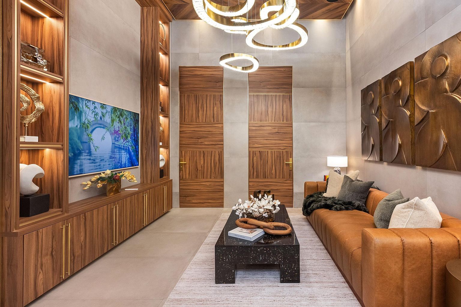 This sophisticated living room features a warm, contemporary aesthetic with rich wood-toned cabinetry and wall paneling. A plush, cognac-colored leather sofa sits opposite a media wall with integrated shelving and a large television, while a dark, textured coffee table serves as a central focal point. The space is illuminated by a striking, multi-ring gold chandelier, and the walls are adorned with three-dimensional bronze-toned art pieces.