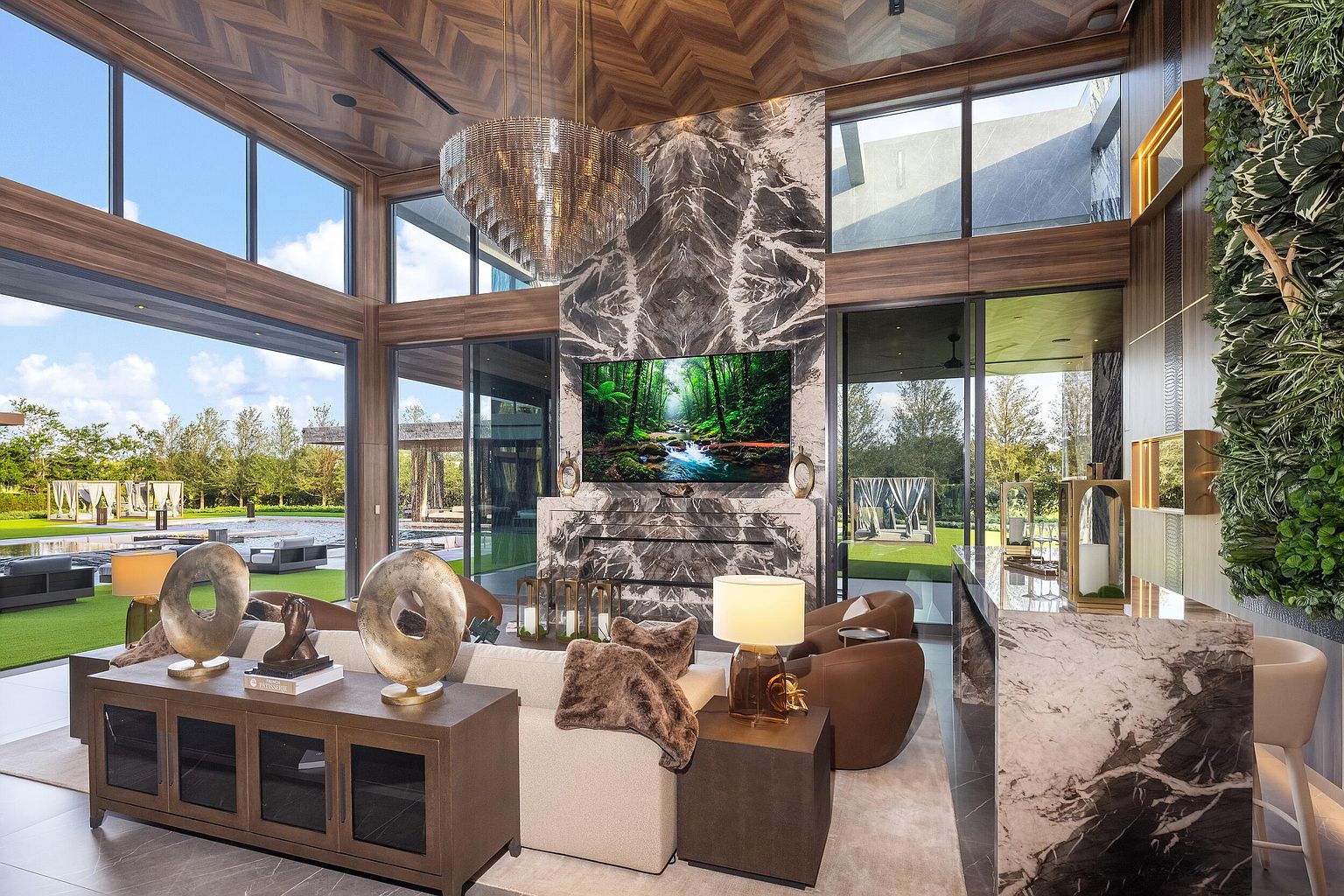 This expansive living room features a dramatic stone-clad chimney breast that serves as a central focal point, complemented by a large television and a sophisticated chandelier. The space is framed by tall windows that offer seamless views of the outdoor pool and manicured grounds, blurring the line between indoor and outdoor living. Rich wood-paneled ceilings and a vertical garden wall add texture and warmth to the contemporary, high-end design.