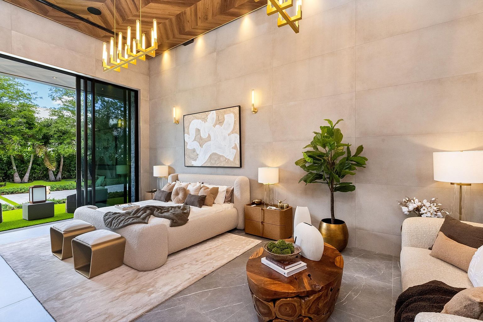 This luxurious primary bedroom features a contemporary design with a textured, neutral-toned bed frame and a chevron-patterned wood ceiling. Large glass sliding doors provide a seamless transition to the outdoor garden, while a natural wood coffee table and plush seating area create a sophisticated lounge space. The room is accented by warm, modern lighting fixtures and large-format wall tiles, offering an elegant and serene atmosphere.