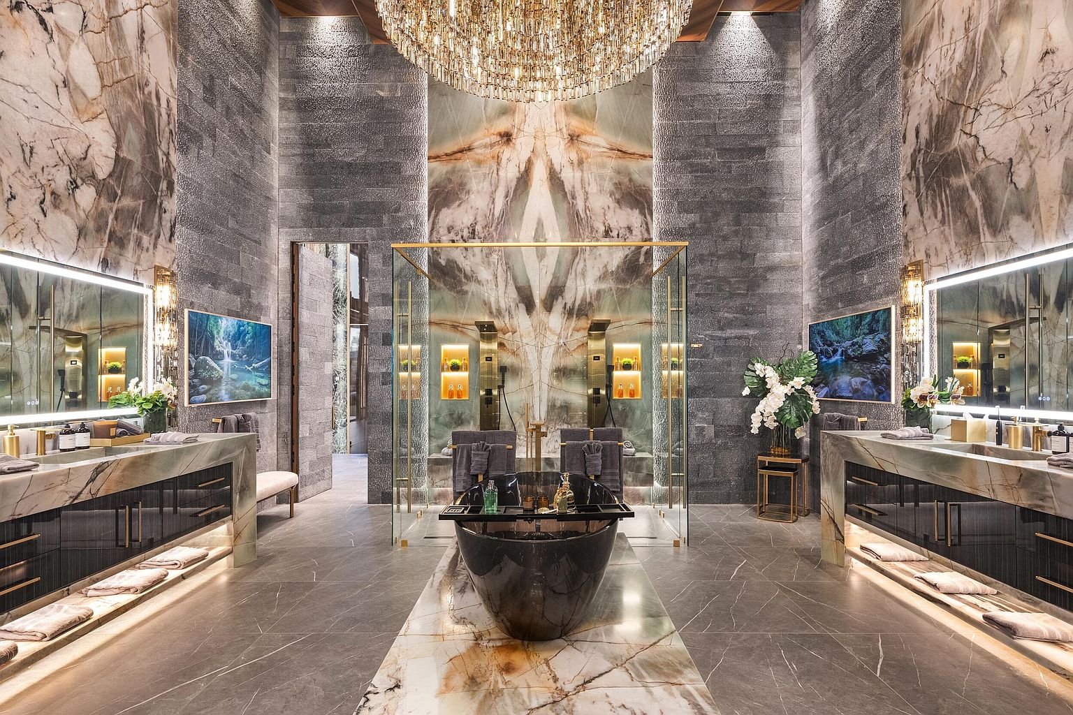 This opulent primary bathroom features a symmetrical layout with dual vanities flanking a central glass-enclosed shower and soaking tub area. The space is defined by dramatic, large-format stone surfaces, a grand crystal chandelier, and integrated ambient lighting that highlights the intricate textures of the walls. The cinematic perspective looks directly down the center axis, emphasizing the luxurious scale and high-end finishes of this spa-like retreat.