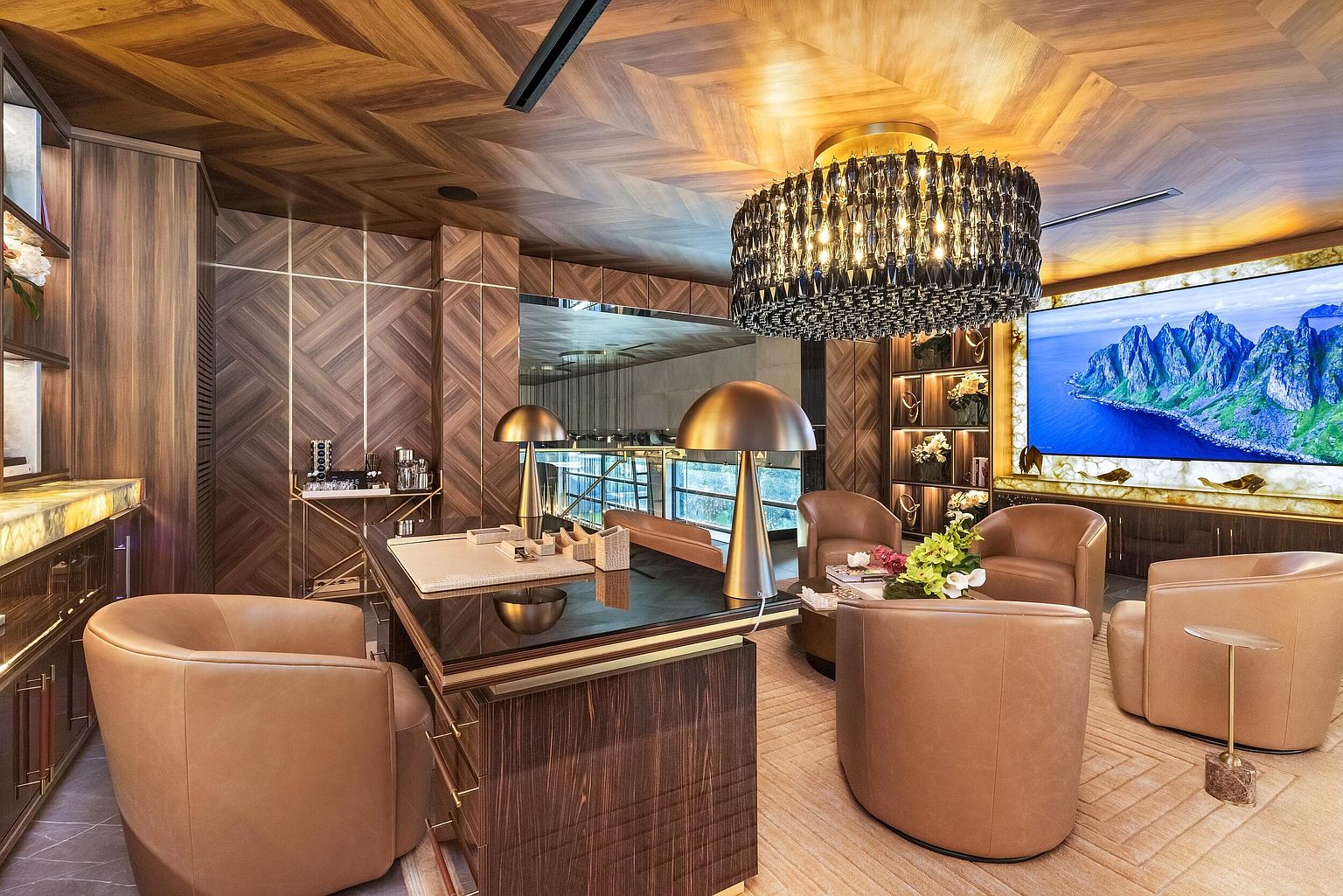 This sophisticated home office features rich, dark wood paneling in a chevron pattern on both the walls and ceiling, creating a cohesive and luxurious atmosphere. The room is anchored by a sleek desk with a dark top and a prominent crystal chandelier, complemented by comfortable tan leather armchairs arranged for conversation. A large wall-mounted television displays a scenic landscape, while integrated shelving and warm, backlit stone accents add depth and elegance to the space.