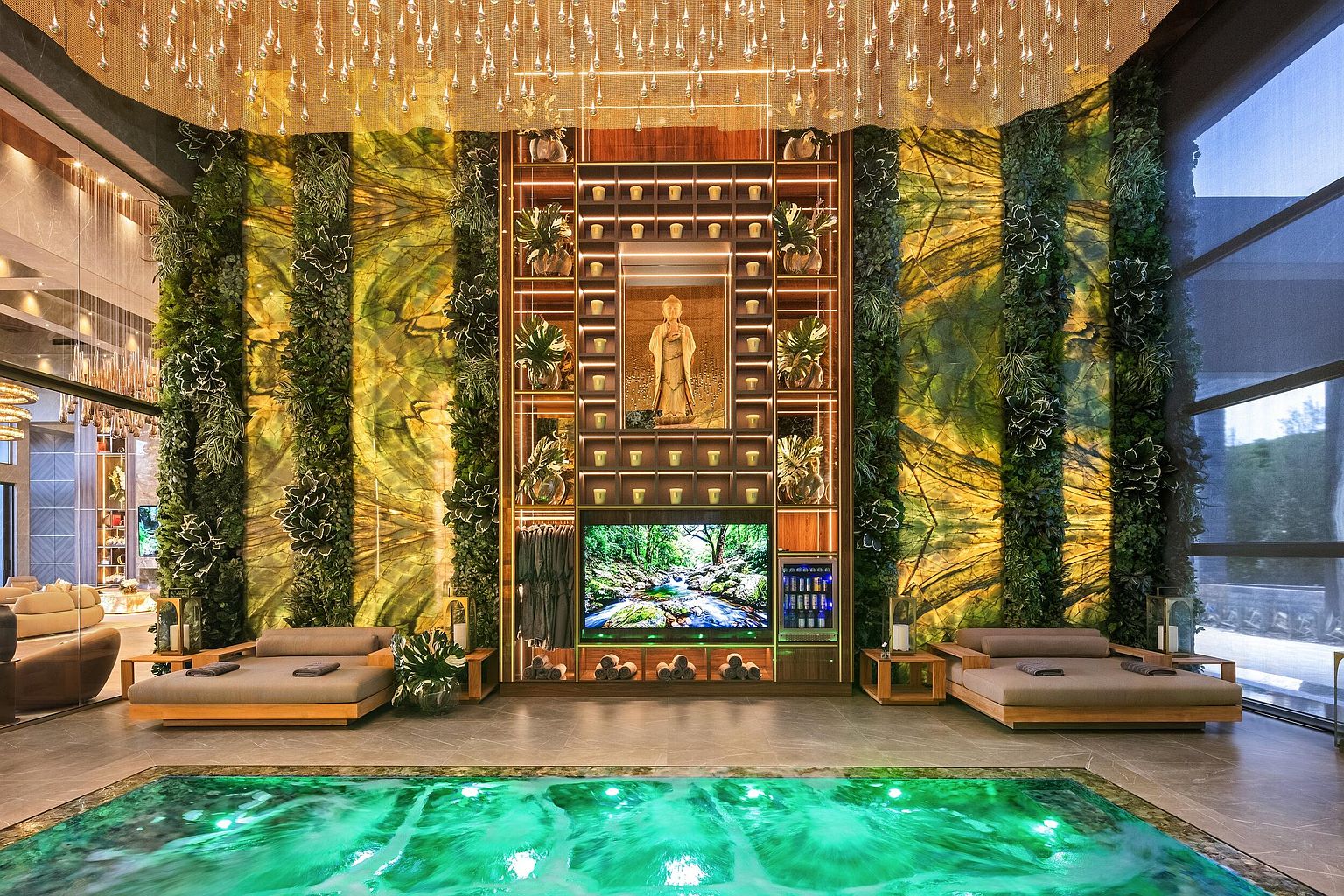 This luxurious indoor spa area features a vibrant, illuminated green pool as the central focal point, set against a dramatic wall adorned with vertical gardens and a central shelving unit housing a Buddha statue. Two plush lounge beds with grey cushions flank the pool, while a large television screen and a built-in beverage cooler are integrated into the central display. The space is crowned by an elaborate, cascading light fixture that creates a sophisticated, tranquil atmosphere suitable for a high-end wellness retreat.