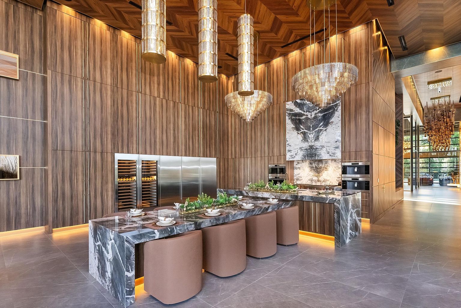 This expansive, high-end kitchen features a massive stone-topped island with integrated seating and a central decorative planter. The space is defined by warm, vertical wood paneling, a dramatic stone-clad chimney breast above the cooking area, and a series of ornate, cylindrical pendant lights. The perspective captures the grandeur of the room, highlighting the seamless integration of professional-grade stainless steel appliances and sophisticated, modern design elements.