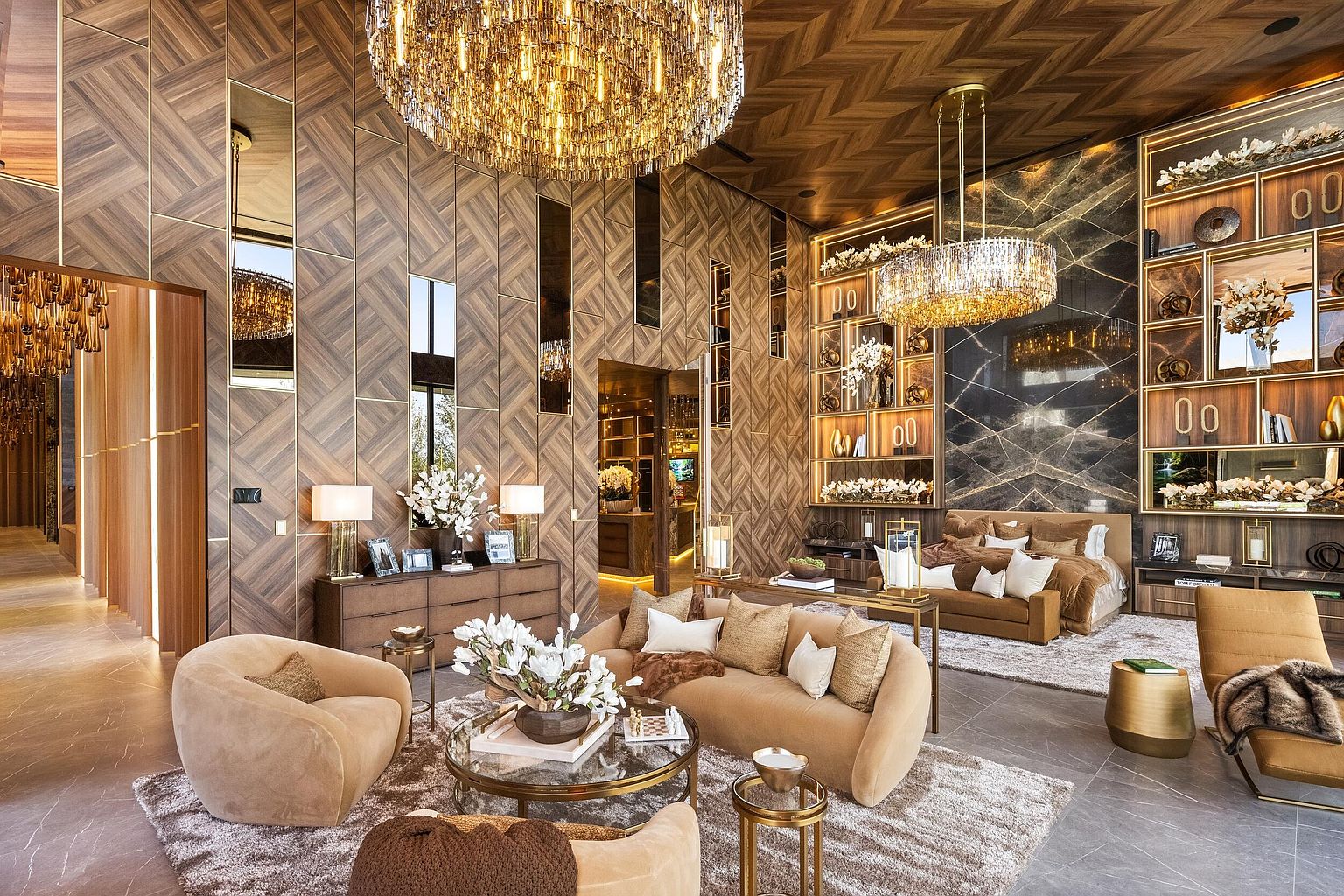 This expansive primary suite features a sophisticated blend of warm wood paneling in a chevron pattern and a dramatic dark stone accent wall behind the bed. The room is illuminated by two large, tiered crystal chandeliers and includes a cozy seating area with plush, neutral-toned furniture arranged around a circular glass coffee table. Custom shelving units integrated into the stone wall provide elegant display space, creating a luxurious and cohesive aesthetic throughout the open-concept layout.