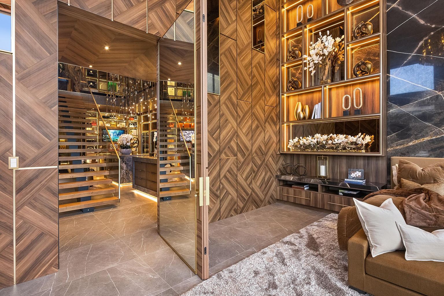 This sophisticated living space features intricate wood-paneled walls with a chevron pattern and a large, illuminated display shelving unit. A glass-paneled door provides a view into an adjacent staircase area, while a plush sofa sits on a textured rug in the foreground. The room is defined by its warm lighting, rich textures, and a seamless blend of modern architectural design and luxury decor.