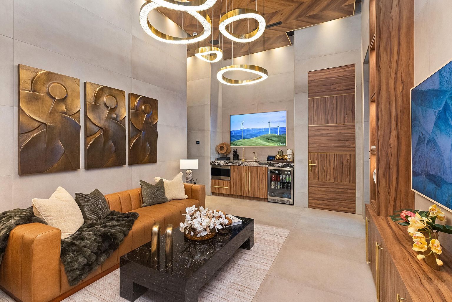 This sophisticated living space features a warm, cognac-colored leather sofa accented with textured throw pillows and a dark, speckled stone coffee table. The room is illuminated by a striking arrangement of circular gold pendant lights suspended from a chevron-patterned wood ceiling, while a wet bar with a built-in beverage cooler and television adds functional luxury. The neutral wall panels and rich wood cabinetry create a refined, contemporary atmosphere perfect for entertaining.