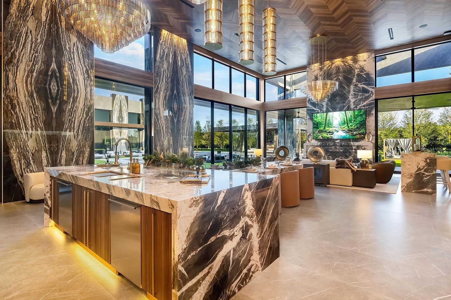 This expansive, open-concept kitchen and living area features a massive central island with a dramatic stone waterfall edge and integrated stainless steel appliances. The space is defined by its soaring ceilings, large windows that provide an unobstructed view of the outdoor landscape, and a striking stone-clad chimney breast behind the living area. The interior design combines warm wood tones with bold, dark stone accents and elegant chandeliers to create a sophisticated, high-end atmosphere.