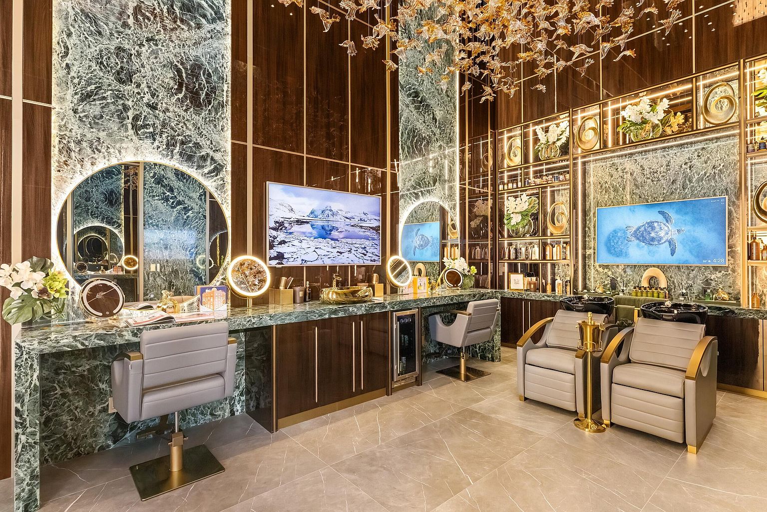 This image showcases a luxurious, high-end salon or spa interior featuring dual styling stations with circular mirrors and grey upholstered chairs. The space is defined by rich, dark wood paneling, green stone surfaces, and gold-toned accents, including a dramatic sculptural chandelier overhead. To the right, two shampoo stations are positioned near a display shelving unit, creating a sophisticated and professional atmosphere.