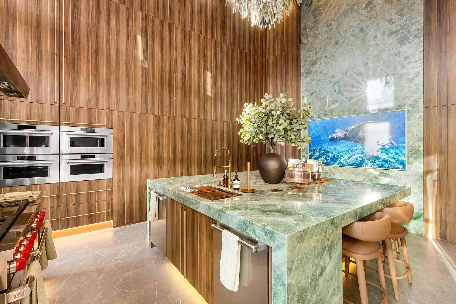 This luxurious kitchen features a striking central island with a green stone waterfall countertop and integrated seating. The space is defined by tall wood-paneled walls, a large wall-mounted television displaying an underwater scene, and high-end stainless steel appliances. A crystal chandelier hangs above, adding a touch of elegance to the sophisticated, modern design.