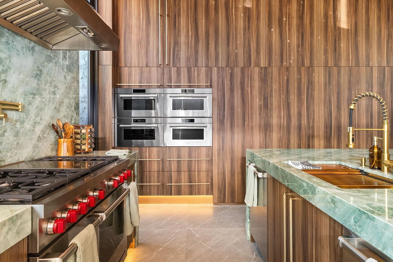This sophisticated kitchen showcases a high-end aesthetic with rich, vertical-grain wood cabinetry and striking green stone countertops. The space is equipped with professional-grade stainless steel appliances, including a large range and a stacked wall oven configuration, complemented by gold-toned hardware and fixtures. The perspective captures the functional workspace, highlighting the seamless integration of luxury materials and modern culinary design.
