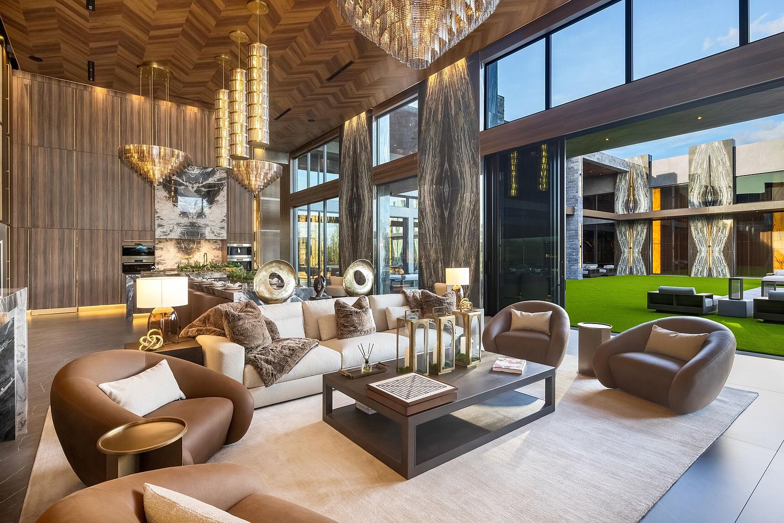 This expansive, open-concept living area features a sophisticated design with a chevron-patterned wood ceiling and large glass walls that open to a manicured courtyard. The space is furnished with a plush, light-toned sofa, curved leather armchairs, and a central coffee table, all arranged on a neutral area rug. A series of tiered, metallic pendant lights hangs above the kitchen area in the background, while the room is anchored by dramatic, dark-toned stone pillars.