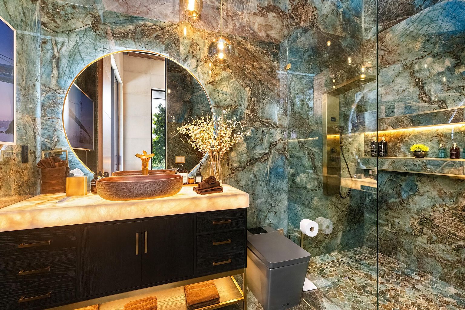 This luxurious bathroom features striking blue-toned stone walls with dramatic veining and a matching hexagonal tile floor. A large, circular mirror hangs above a floating vanity with a backlit stone countertop and a textured, oval-shaped vessel sink. To the right, a glass-enclosed shower area includes built-in shelving with integrated lighting, creating a sophisticated and high-end spa-like atmosphere.