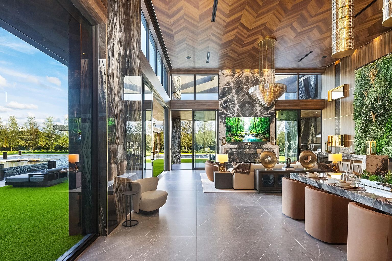This expansive, open-concept living area features a dramatic stone-clad chimney breast that serves as a focal point, complemented by a herringbone-patterned wood ceiling. Large glass walls provide a seamless transition to the outdoor patio and pool area, while a lush vertical garden wall adds a vibrant organic element to the space. The room is furnished with contemporary seating, a long dining table, and a sophisticated crystal chandelier, creating an atmosphere of refined luxury.
