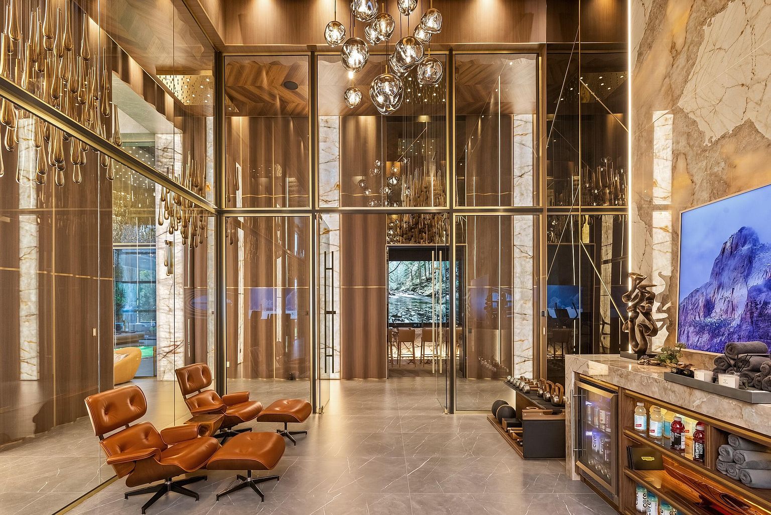 This sophisticated home gym features a sleek, modern aesthetic with dark stone flooring and expansive glass partitions that create an open, airy feel. The space is furnished with two iconic lounge chairs and ottomans, a beverage refrigerator, and a selection of hand weights, all illuminated by a striking cluster of spherical pendant lights. The room offers a seamless transition into an adjacent area, emphasizing a luxurious, high-end design suitable for a private wellness retreat.