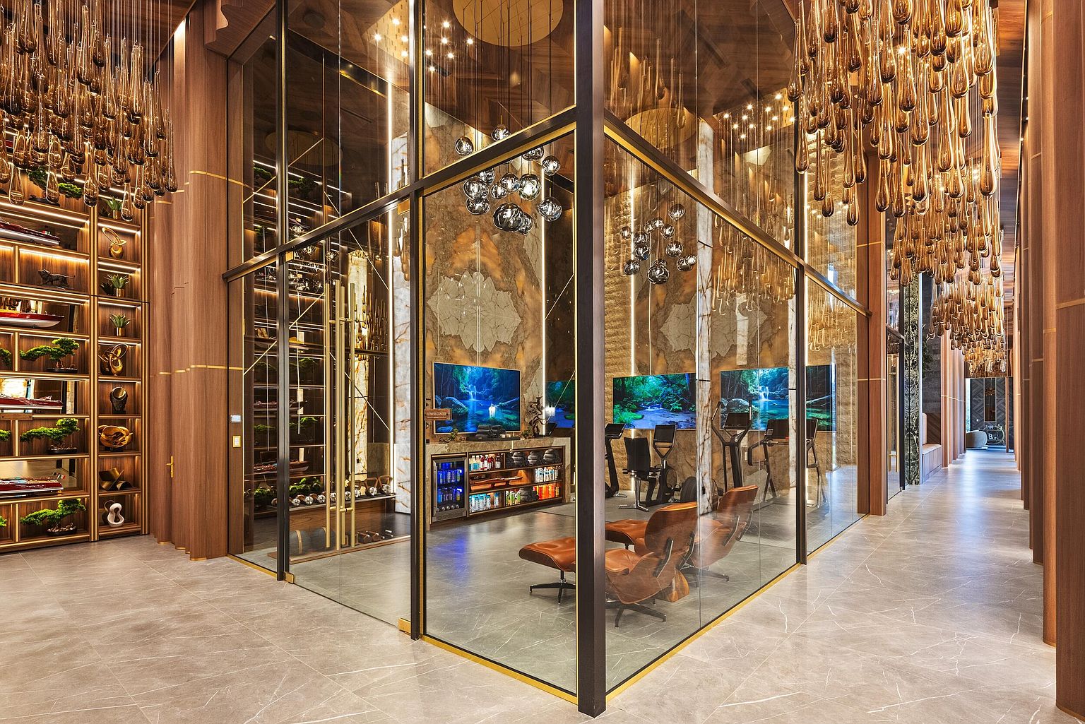 This sophisticated home gym is enclosed within a glass-walled structure, featuring a sleek, modern aesthetic with warm wood paneling and stone flooring. The space is equipped with exercise equipment, lounge chairs, and a beverage refrigerator, all illuminated by dramatic, cascading light fixtures. The perspective looks into the gym from a hallway, highlighting the high-end finishes and the seamless integration of luxury design.