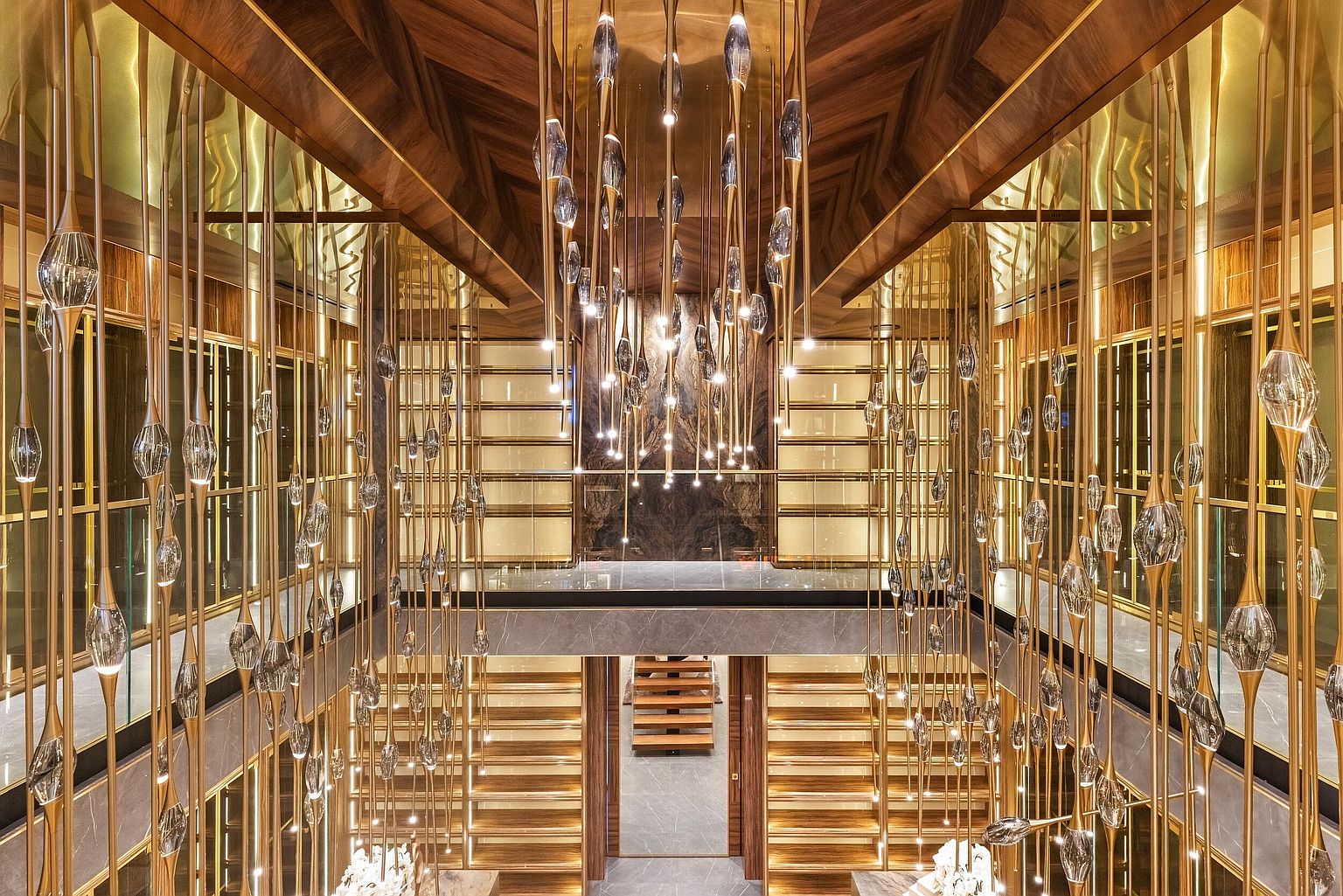 This dramatic interior view captures a grand staircase and landing, defined by a massive, multi-tiered chandelier featuring numerous glass pendants suspended on slender gold rods. The space is characterized by rich wood paneling, gold-toned metal accents, and polished stone surfaces that create a luxurious, high-end atmosphere. The perspective looks down from an upper level, emphasizing the verticality and intricate lighting design of this opulent architectural feature.