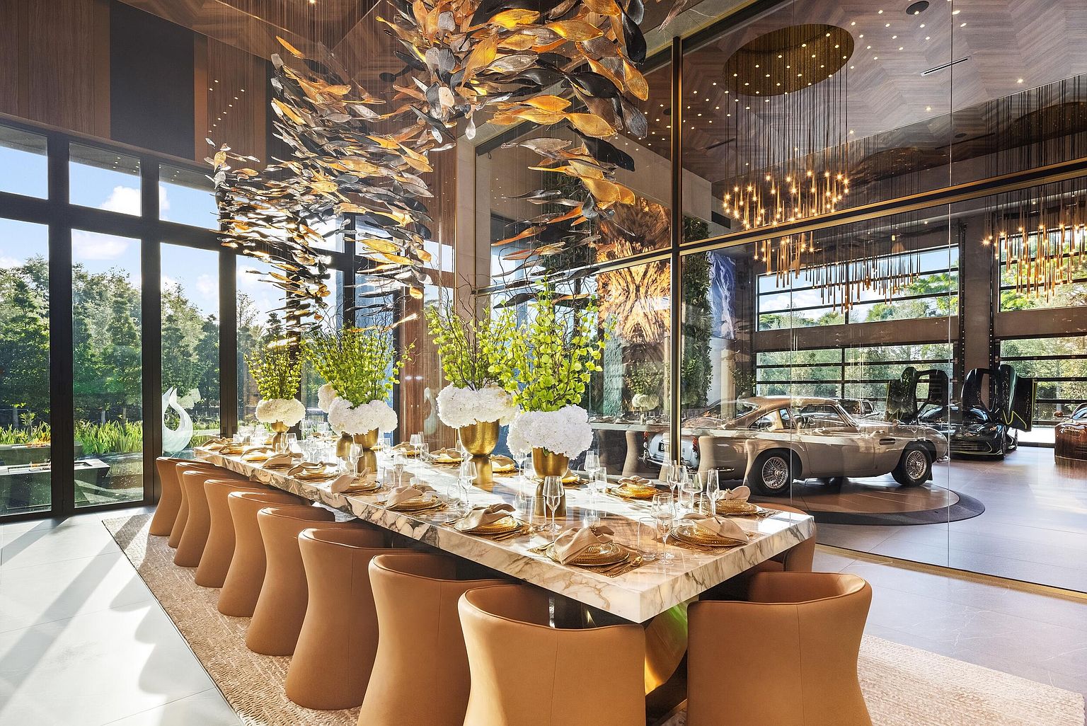 This sophisticated dining room features a long, stone-topped table surrounded by tan leather chairs, set beneath a dramatic, sculptural metallic light fixture. Large windows provide an expansive view of the lush greenery outside, while a glass wall separates the dining area from a gallery-style garage showcasing luxury vehicles. The space exudes a high-end, modern aesthetic with a seamless blend of natural light and refined interior design.