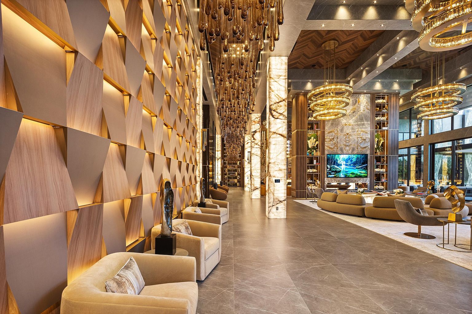 This expansive, high-end living area features a dramatic, multi-layered lighting design with cascading glass pendants and circular chandeliers. The space is defined by a striking geometric wood-paneled wall with integrated lighting and a series of plush, neutral-toned armchairs arranged along a polished stone floor. A large television is mounted on a stone-clad wall, flanked by illuminated shelving units, creating a sophisticated and luxurious atmosphere suitable for a grand lobby or estate lounge.