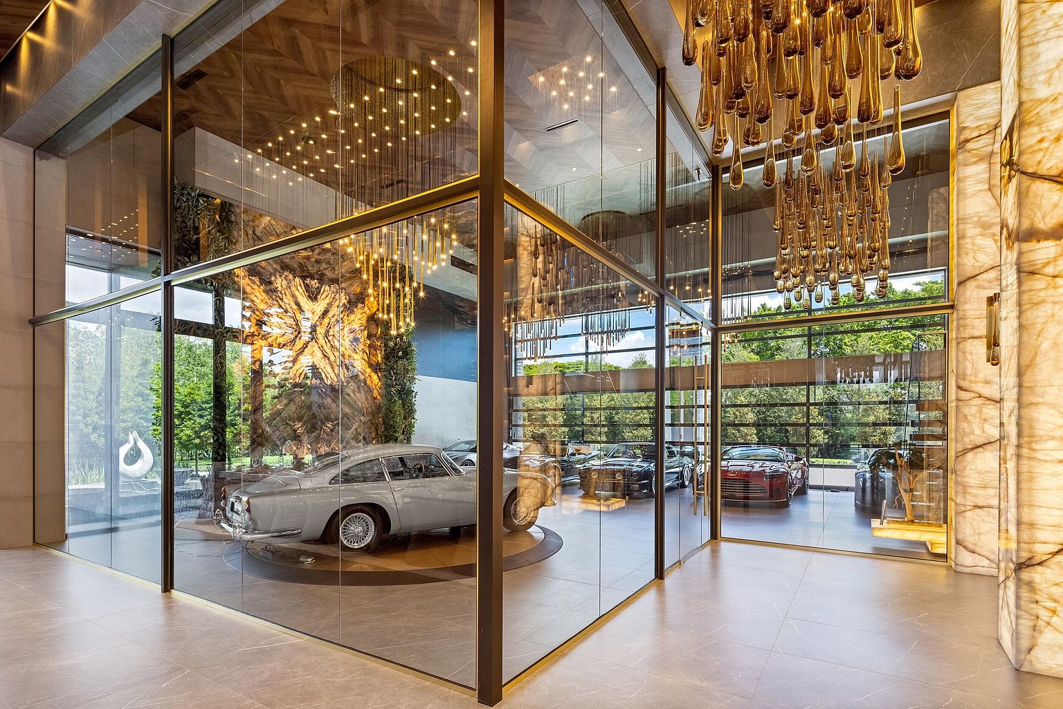 This sophisticated interior space features a glass-enclosed display area showcasing a collection of luxury vintage and modern automobiles. The room is accented by dramatic, cascading gold-toned pendant lighting and a striking, illuminated stone feature wall that serves as a focal point. The perspective captures the sleek, modern aesthetic of a high-end gallery or private showroom, emphasizing the clean lines of the glass partitions and the polished tile flooring.