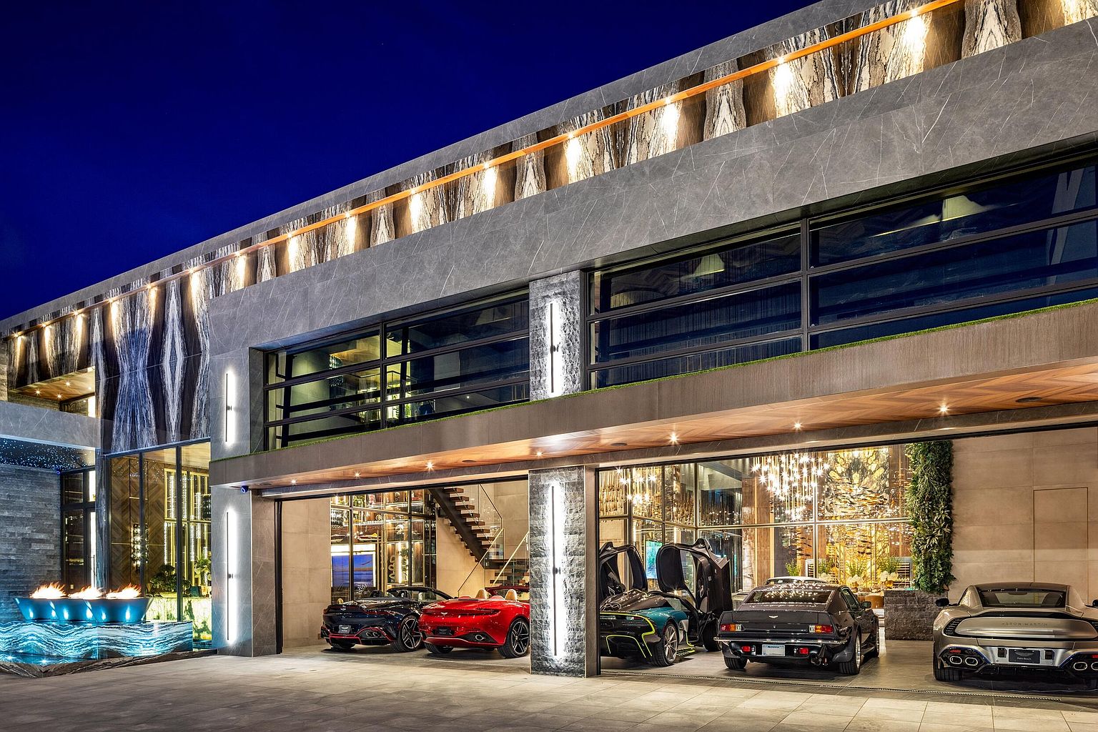 This striking modern garage features a multi-bay design with large glass-paneled doors that showcase a collection of luxury vehicles. The exterior is clad in textured stone panels with integrated linear lighting, while a water feature with a fire element adds a dramatic touch to the foreground. The overall aesthetic is ultra-contemporary and high-end, emphasizing a seamless blend of architectural design and automotive display.
