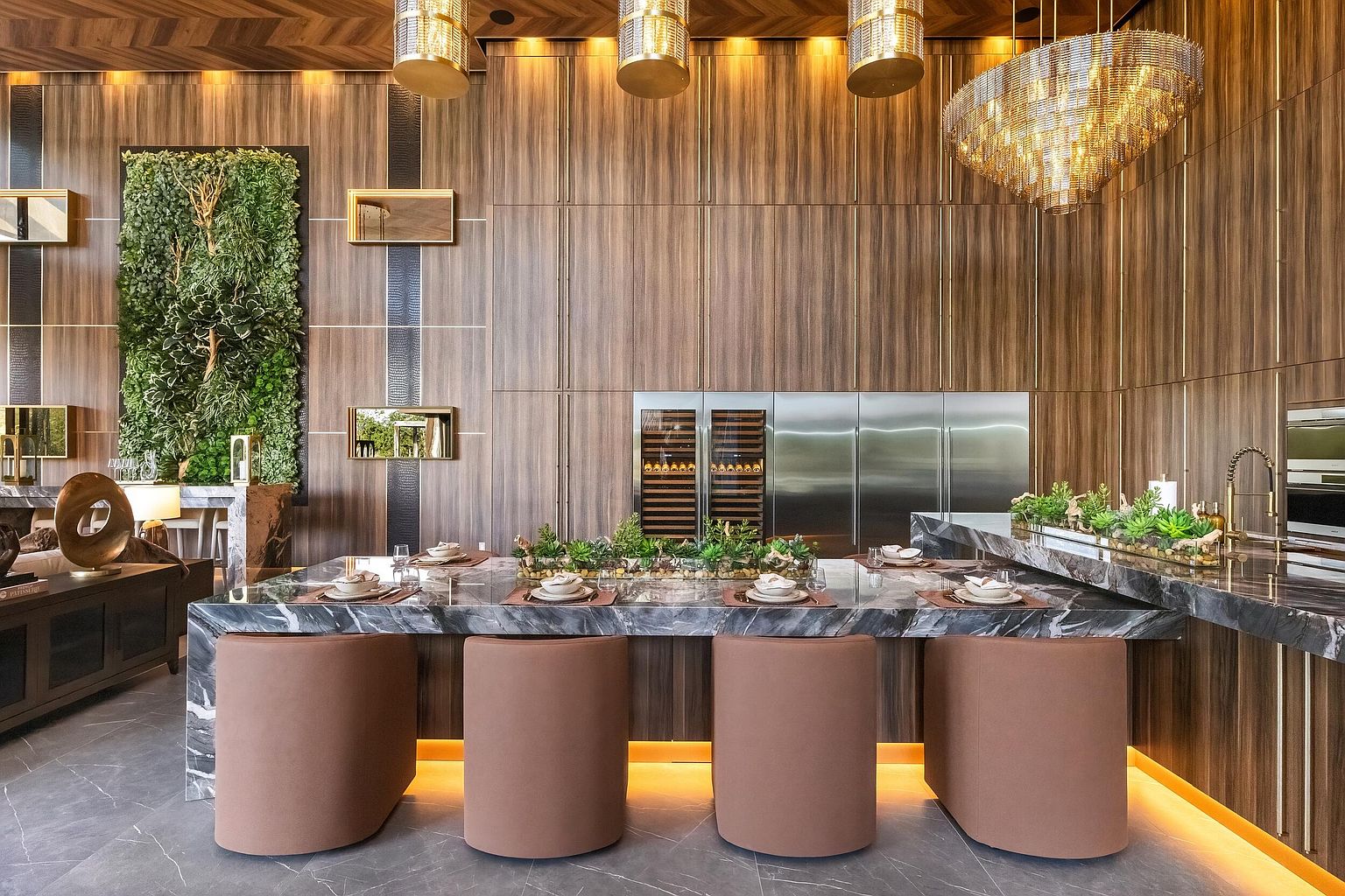 This sophisticated, high-end kitchen features a large stone-topped island with integrated seating and a central succulent planter, illuminated by warm under-cabinet lighting. The space is defined by rich, vertical wood-paneled walls, a striking vertical garden feature, and a dramatic crystal chandelier that adds a touch of luxury. The perspective is centered on the island, showcasing a seamless blend of natural textures and modern, professional-grade appliances.
