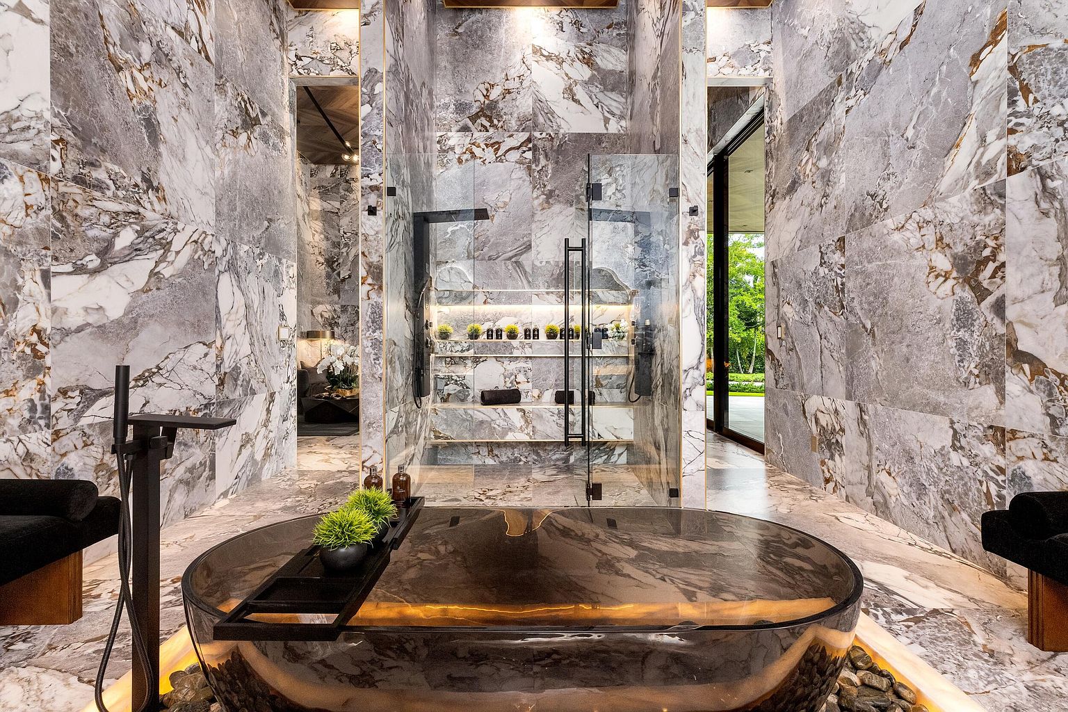 This luxurious primary bathroom features a striking, dark-toned soaking tub as the centerpiece, accented by a wooden tray with greenery. The space is clad in dramatic, large-format patterned stone tiles that extend across the walls and floor, creating a cohesive and opulent aesthetic. A glass-enclosed shower with integrated shelving and warm accent lighting sits between two doorways, while the overall design emphasizes a high-end, spa-like atmosphere.