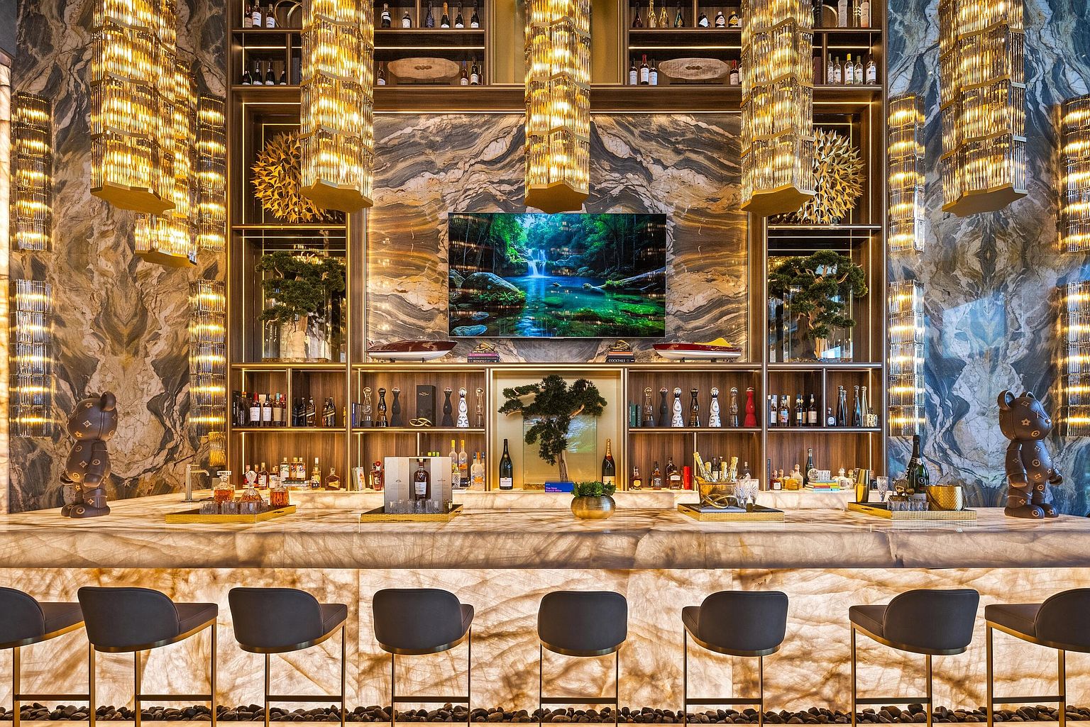 This image showcases a luxurious, high-end home bar area characterized by a backlit stone countertop and matching stone wall cladding. The space is defined by symmetrical gold-toned pendant lighting, open shelving filled with premium spirits, and two decorative bear figurines flanking the bar. The cinematic perspective is centered and balanced, emphasizing the opulent materials and the integrated television displaying a vibrant waterfall scene.