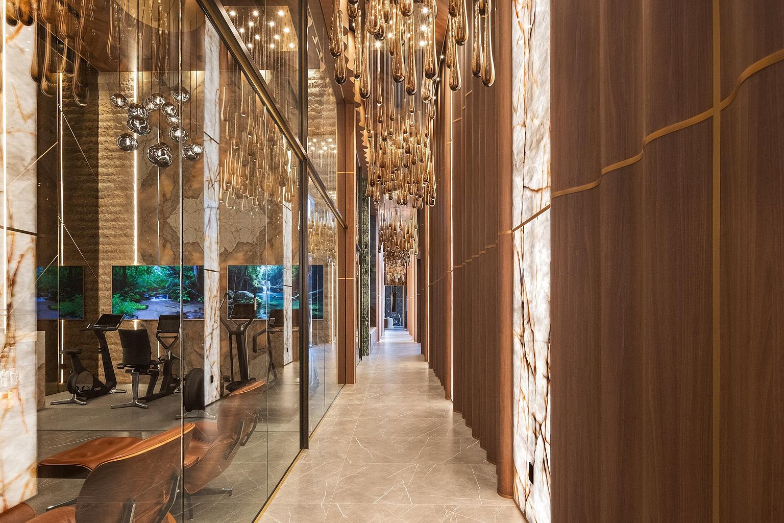 This sophisticated hallway features a long corridor lined with warm-toned wood paneling accented by vertical gold trim and illuminated stone panels. To the left, a glass partition reveals a modern home gym equipped with exercise machines and screens displaying nature scenes, while a series of elegant, teardrop-shaped pendant lights hang from the ceiling, creating a dramatic, high-end atmosphere. The perspective draws the eye down the length of the hall, emphasizing the luxurious materials and refined architectural design.