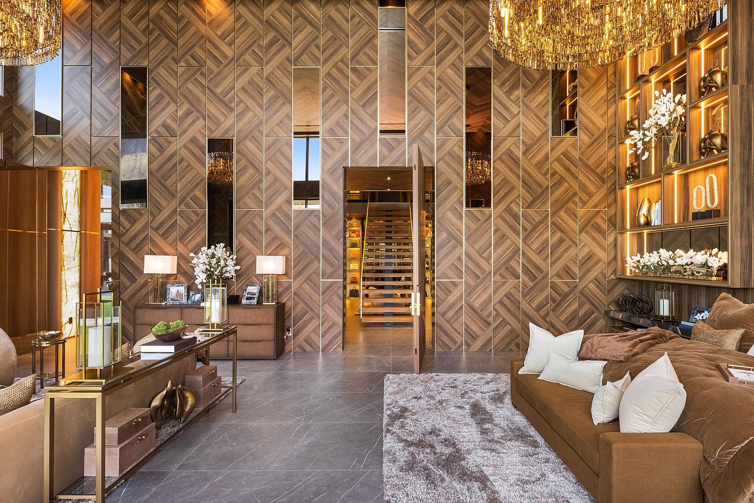 This opulent living space features a dramatic wall clad in a herringbone wood pattern, accented by vertical mirror panels and integrated gold trim. A plush brown sofa sits on a textured rug, while a sleek console table with gold accents and a large crystal chandelier add to the sophisticated, high-end aesthetic. The room opens into a central doorway revealing a staircase, creating a sense of depth and architectural grandeur.