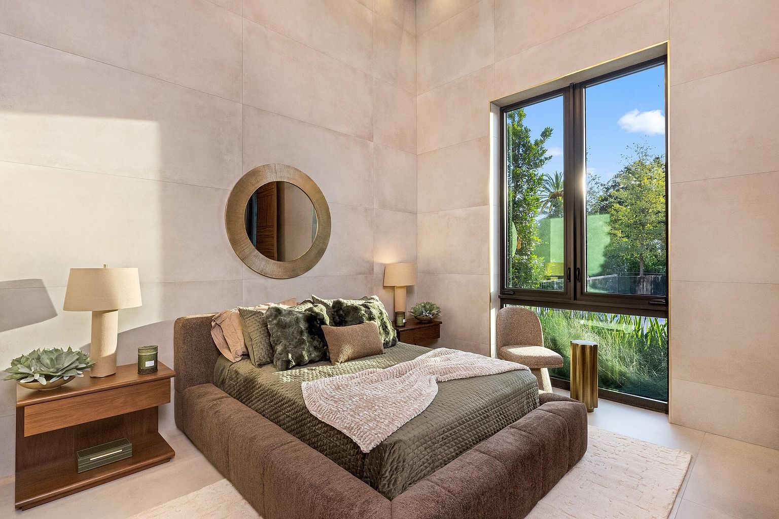 This modern bedroom features a low-profile, upholstered bed frame centered against a wall finished with large, neutral-toned panels. A circular mirror hangs above the bed, while a tall window to the right provides ample natural light and a view of the lush greenery outside. The room is accented by a wooden nightstand with a lamp and a small, textured chair, creating a serene and contemporary atmosphere.