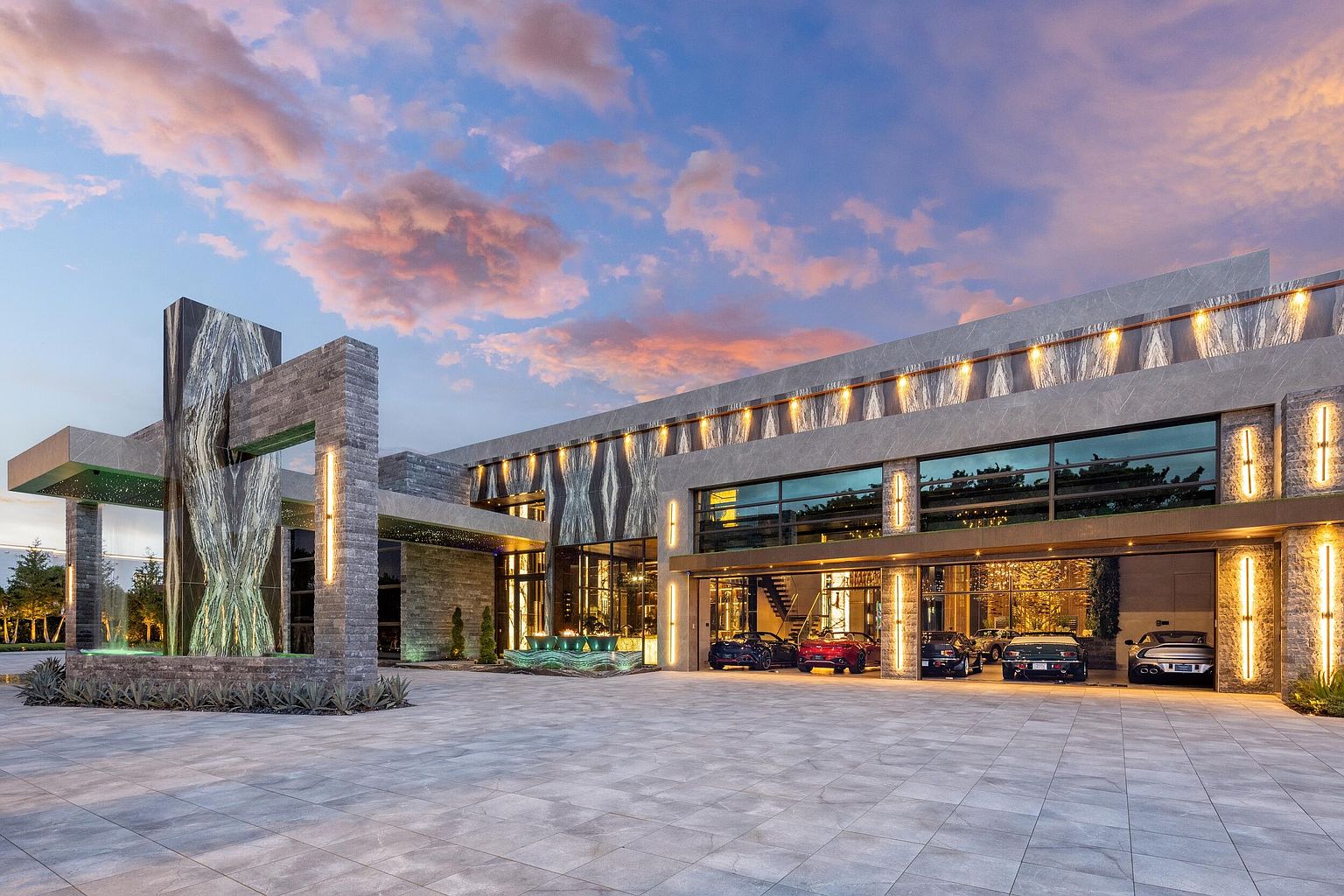 This striking modern estate features a dramatic, multi-level facade characterized by large stone-clad pillars and expansive glass walls that showcase an impressive collection of luxury vehicles. The architectural design incorporates a prominent water feature at the entrance and warm, integrated lighting that highlights the textured stone surfaces against a vibrant sunset sky. The wide, paved motor court provides a grand sense of arrival, emphasizing the property's scale and contemporary luxury aesthetic.
