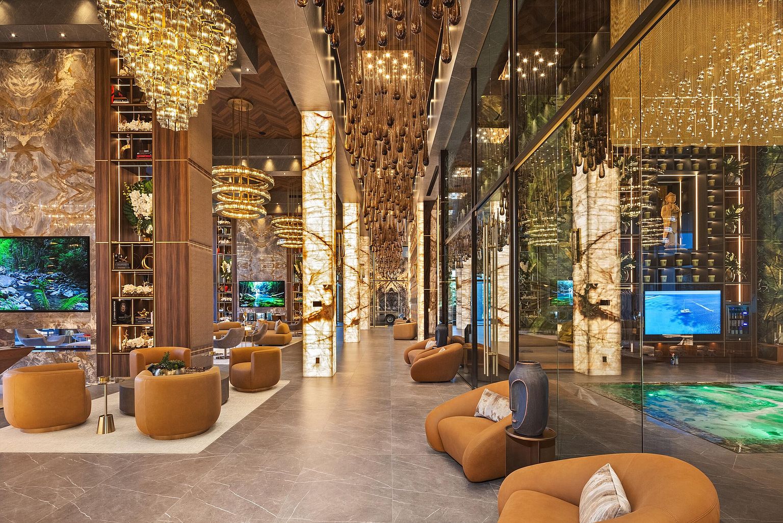 This opulent lobby or lounge area features a sophisticated design with warm, ambient lighting, numerous tan-colored armchairs, and striking illuminated stone pillars. The space is adorned with elaborate, cascading crystal chandeliers and large glass partitions that reveal a vibrant, illuminated indoor water feature. The overall aesthetic is one of high-end luxury, characterized by rich textures, dark wood accents, and a spacious, open-concept layout.